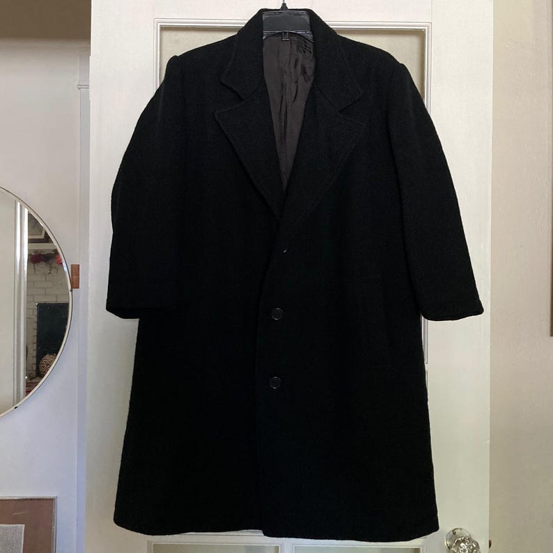 May include: A solid black long-sleeve overcoat featuring a notched lapel collar, a straight hemline, and a front button closure with two visible dark buttons. The garment is fully lined with a dark brown interior fabric and hangs on a black hanger.