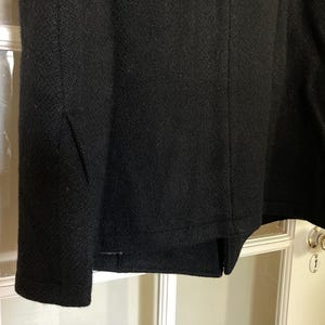May include: A black coat featuring a textured fabric surface with a visible vertical seam running down the center and a side panel detail. The garment has a straight hemline and a consistent dark color throughout the visible portion of the fabric.