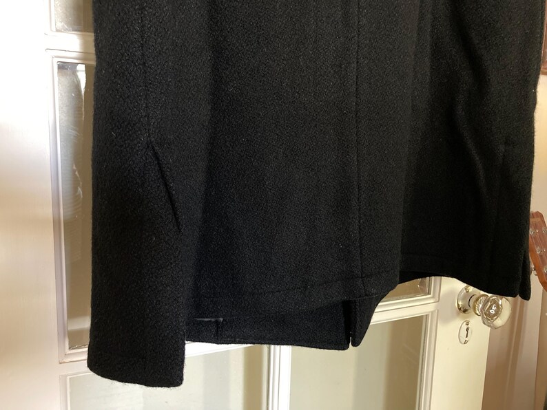 May include: A black coat featuring a textured fabric surface with a visible vertical seam running down the center and a side panel detail. The garment has a straight hemline and a consistent dark color throughout the visible portion of the fabric.