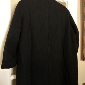 May include: A long, black coat featuring a structured collar, long sleeves, and a central vertical seam running down the back. The garment is made of a textured, dark fabric and is shown hanging from a hanger, displaying a simple, clean silhouette.