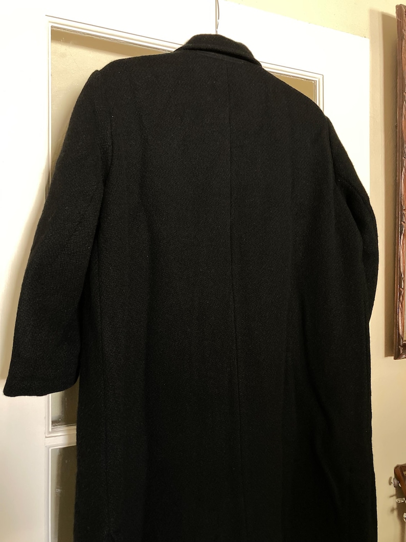May include: A long, black coat featuring a structured collar, long sleeves, and a central vertical seam running down the back. The garment is made of a textured, dark fabric and is shown hanging from a hanger, displaying a simple, clean silhouette.