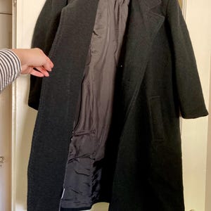 May include: A long, dark charcoal gray coat featuring a structured collar, long sleeves, and two side pockets. The coat is open at the front, revealing a smooth, dark interior lining. The outer fabric has a textured, wool-like appearance.