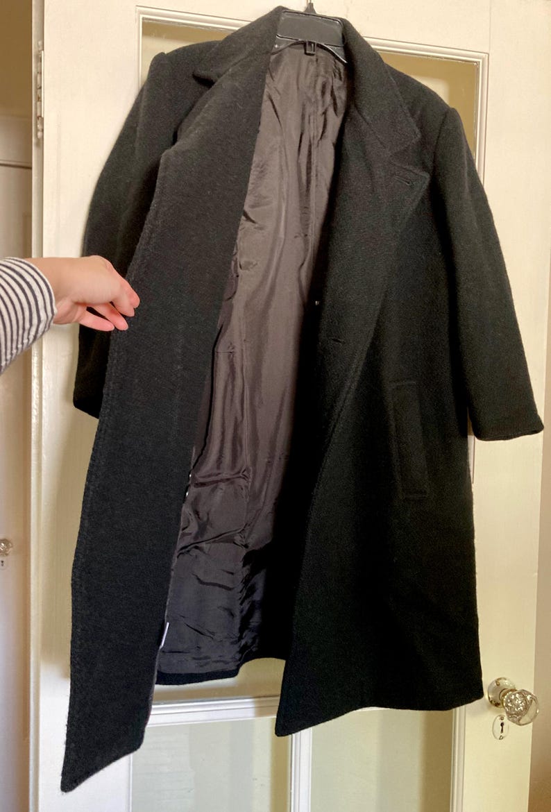 May include: A long, dark charcoal gray coat featuring a structured collar, long sleeves, and two side pockets. The coat is open at the front, revealing a smooth, dark interior lining. The outer fabric has a textured, wool-like appearance.