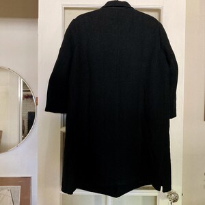 May include: A solid black long-length coat featuring a structured collar, long sleeves, and a straight hemline. The garment is displayed from the back, showcasing a clean, unadorned design with a central vertical seam running down the back panel.