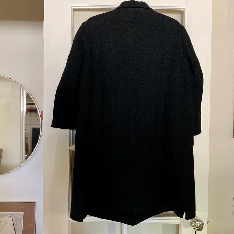May include: A solid black long-length coat featuring a structured collar, long sleeves, and a straight hemline. The garment is displayed from the back, showcasing a clean, unadorned design with a central vertical seam running down the back panel.
