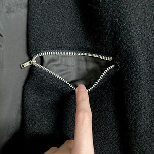 May include: A black textured fabric garment featuring a diagonal zippered pocket. The silver-toned metal zipper is partially open, revealing a smooth black interior lining. The fabric has a dense, slightly coarse weave characteristic of wool.