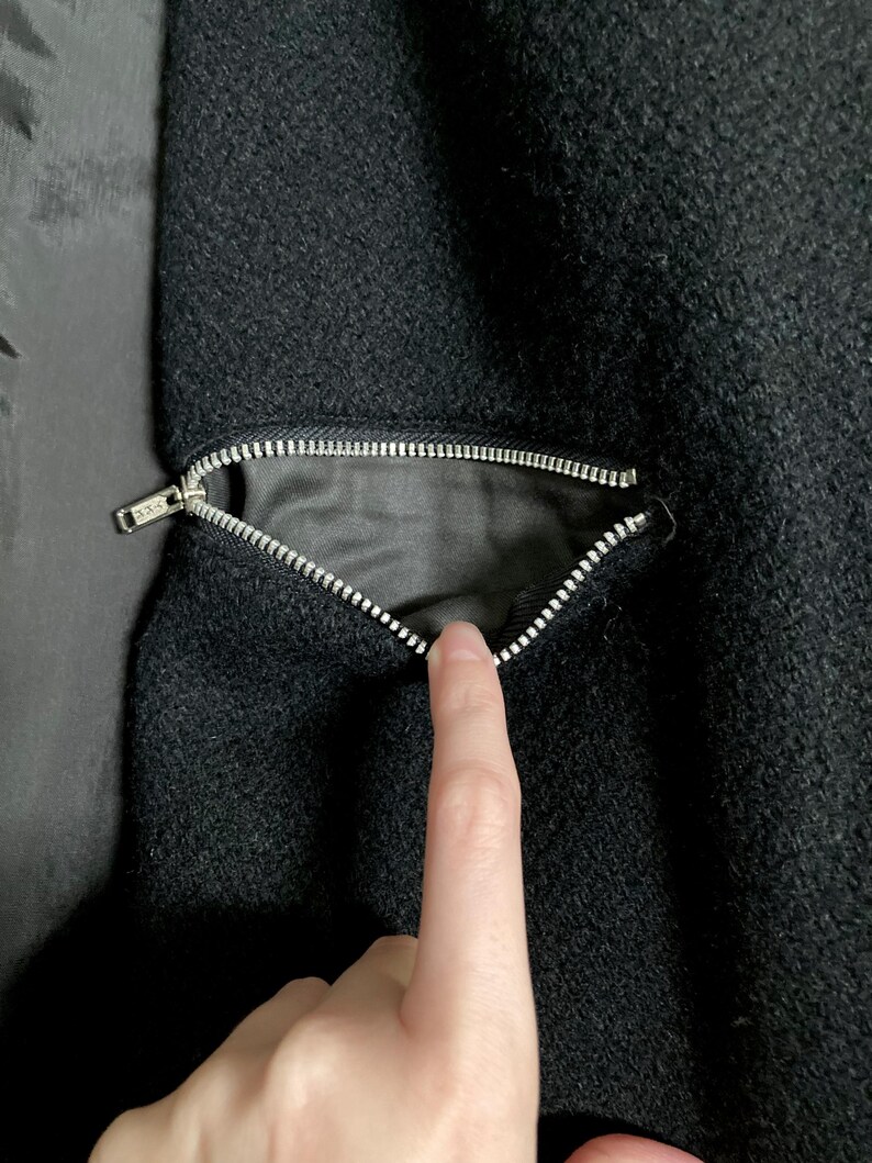 May include: A black textured fabric garment featuring a diagonal zippered pocket. The silver-toned metal zipper is partially open, revealing a smooth black interior lining. The fabric has a dense, slightly coarse weave characteristic of wool.