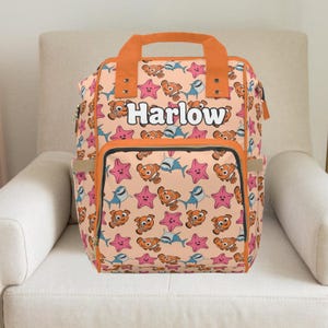 May include: A peach-colored backpack with an orange handle and trim, featuring a pattern of cartoon fish, starfish, and sharks. The name "Harlow" is printed in white on the front. The bag has multiple pockets and a zipper closure.
