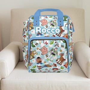 Peut inclure: Un sac à dos bleu clair et blanc avec des personnages de Toy Story. Le sac a une poignée et une bordure bleues, avec le nom "Rocco" imprimé sur le devant. Le sac est posé sur une chaise crème. Hauteur d'environ 40 cm.