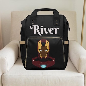 Pu&ograve; includere: Borsa per pannolini nera con una cerniera dorata e un design di Iron Man rosso e oro. La borsa ha il nome "River" stampato sul davanti.