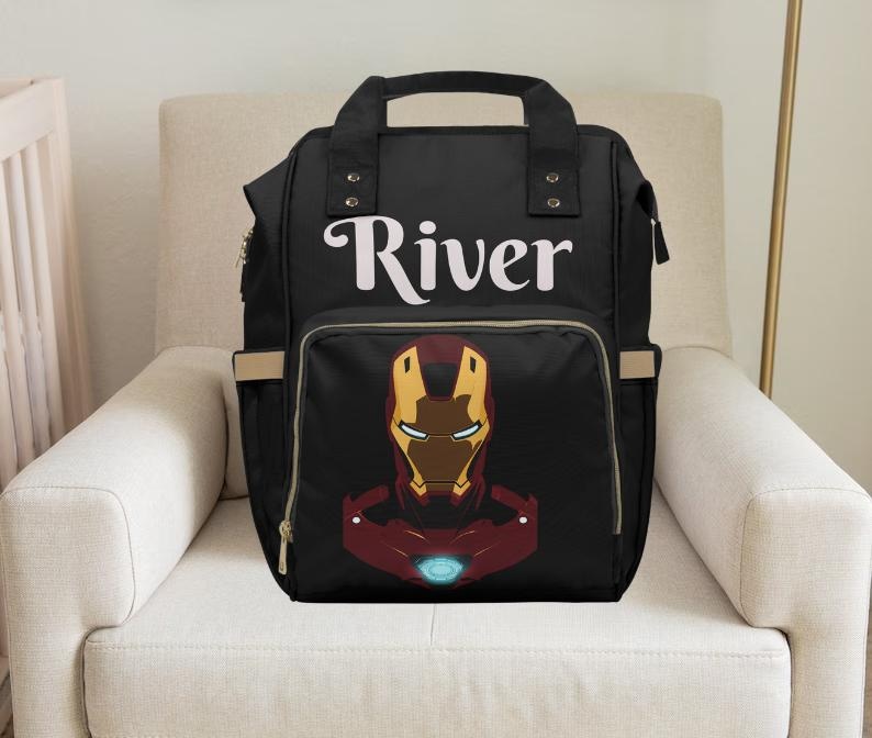 Pu&ograve; includere: Borsa per pannolini nera con una cerniera dorata e un design di Iron Man rosso e oro. La borsa ha il nome "River" stampato sul davanti.