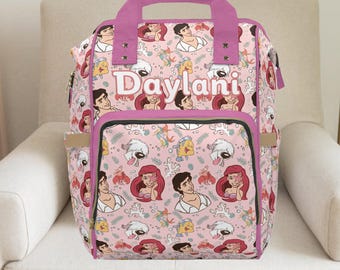 Puede incluir: Mochila rosa con un estampado repetido de personajes de dibujos animados, incluyendo una sirena y un príncipe. La mochila tiene un asa y ribetes morados, con el nombre "Daylani" impreso en la parte delantera.