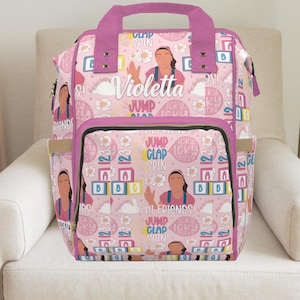 Peut inclure: Sac à langer rose et violet avec un motif répété d'un personnage de dessin animé, de fleurs et de blocs. Le sac a plusieurs poches, une poignée supérieure et le nom "Violetta" en écriture.
