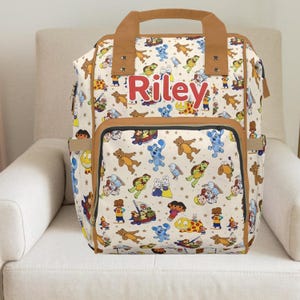 May include: Cream-coloured backpack with a repeating pattern of cartoon characters. The backpack has brown trim, a brown handle, and the name "Riley" in red letters. Features multiple pockets and a front flap.