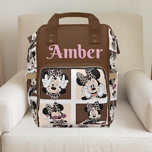 The bag has a brown base color with a design featuring multiple faces of Minnie Mouse, each wearing a cheetah-print bow. The Minnie Mouse faces are displayed in square panels across the front pocket and sides of the bag, showing different expressions such as smiling, winking, and wearing pink sunglasses. At the top of the bag, above the front pocket, the name "Amber" is printed in large, light pink letters. although it says amber you can put any name