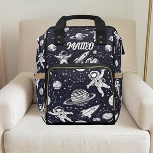 May include: A black diaper bag with a space theme print. The bag features astronauts, planets, stars, and the name "MATTEO" in white lettering.