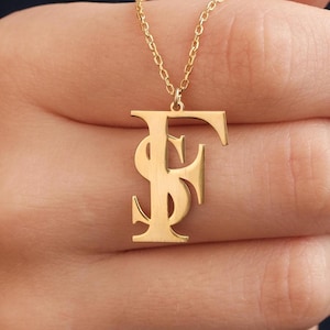以下が含まれることがあります： 「F」と「S」の文字が絡み合ったペンダントが付いたゴールドネックレス。ペンダントはクラシックで太字のフォントです。