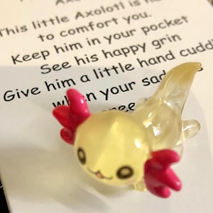 May include: A small, translucent yellow axolotl figurine with bright pink external gills and a smiling face, positioned in front of a white card featuring the text: Pocket Comfort Axolotl, This little Axolotl is here to comfort you.
