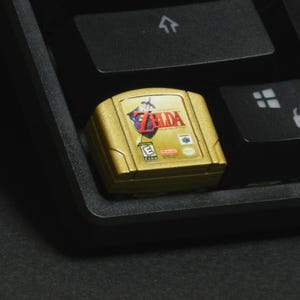 May include: A gold-coloured Nintendo 64 game cartridge for The Legend of Zelda, on a black keyboard. The cartridge displays the game's logo in red and white, with a green 'E' rating symbol.