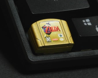 May include: A gold-colored Nintendo 64 game cartridge for The Legend of Zelda, resting on a black keyboard. The cartridge features the game's logo in red and white, with a green 'E' rating symbol.