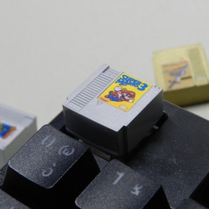 Op de afbeelding: Een close-up van een zwart toetsenbord met drie retro videogame cartridges. De Super Mario Bros. 3 cartridge staat scherp, met een kleurrijk label. Andere cartridges zijn zichtbaar op de achtergrond.
