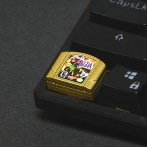 Peut inclure: Une cartouche de jeu vidéo Nintendo 64 dorée pour "The Legend of Zelda" est posée sur un clavier noir. La cartouche présente des illustrations colorées et le titre du jeu. Les touches du clavier sont noires avec des lettres blanches.