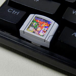 Può includere: Una cartuccia grigia per videogiochi Nintendo 64 con il gioco "Banjo-Kazooie" su una tastiera nera. L'etichetta del gioco è colorata, con personaggi dei cartoni animati e il titolo del gioco. I tasti della tastiera sono neri con scritte bianche.
