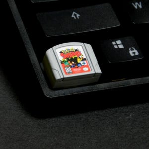 May include: A gray Nintendo 64 video game cartridge for the game "Pokemon Snap" rests on a black keyboard. The cartridge features a colorful label with the game's title and various characters.