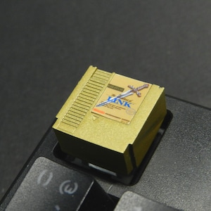 Puede incluir: Una tecla dorada con un diseño de cartucho de videojuego retro. La etiqueta muestra el título "The Legend of Zelda" con una ilustración de espada. La tecla rectangular está sobre un teclado negro.