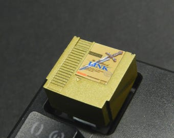 May include: A gold-coloured keycap with a retro video game cartridge design. The label shows "The Legend of Zelda" with a sword illustration. The rectangular keycap is on a black keyboard.