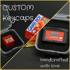May include: Custom keycaps in clear plastic cases, featuring miniature versions of a red Game Boy cartridge and a Pokemon Mystery Dungeon Red Rescue Team game cartridge. The text "CUSTOM Keycaps" is at the top of the image, and the text "handcrafted with love" is at the bottom of the image.