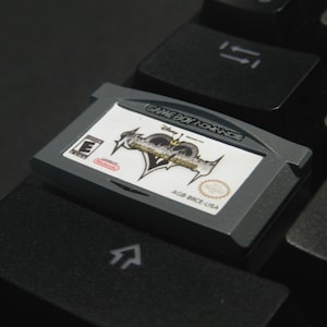 May include: A grey Game Boy Advance cartridge for "Kingdom Hearts: Chain of Memories" sits on a black keyboard. The cartridge has a white label with game details and the Nintendo logo.