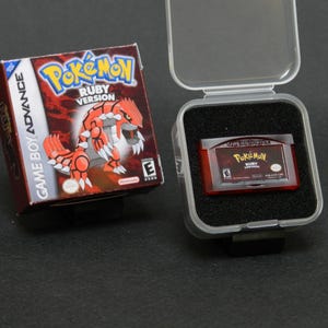 May include: A Game Boy Advance Pokémon Ruby Version game cartridge and box. The box is red and features a graphic of a red and white creature. The cartridge is in a clear plastic case with black foam.