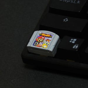 Peut inclure: Une cartouche de jeu grise Nintendo 64 avec le jeu "Super Smash Bros." avec Mario et d'autres personnages. La cartouche est posée sur un clavier noir.