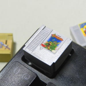 Può includere: Una cartuccia di gioco Nintendo argentata con l'etichetta "Castlevania" è inserita in una console di gioco nera. Sono visibili altre due cartucce di gioco: una gialla e una grigia, con un'etichetta colorata. Le cartucce sono probabilmente per un sistema di gioco retrò.