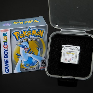 May include: A silver Game Boy Color cartridge with a Pokemon Silver game case. The case features a blue and white Pokemon character with the text "Gotta catch 'em all!" and "Silver Version".