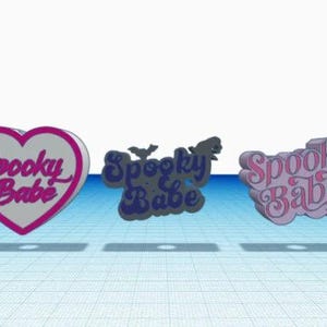 Może przedstawiać: Trzy różne wzory zawieszki "Spooky Babe". Pierwsza zawieszka to różowe serce z białym tłem i napisem "Spooky Babe" w kolorze różowym. Druga zawieszka to wzór w kolorze niebieskim i białym z napisem "Spooky Babe" w kolorze niebieskim. Trzecia zawieszka to wzór w kolorze różowym i białym z napisem "Spooky Babe" w kolorze różowym.