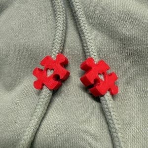 May include: Two red plastic puzzle-shaped toggles with a heart cutout in the centre, attached to a grey hoodie drawstring.