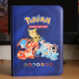 May include: A blue Pokémon Trading Card Game binder featuring the yellow Pikachu, orange Charizard, and blue Blastoise characters on the front cover. The cover includes the Pokémon logo, the text Trading Card Game, and various elemental symbols.