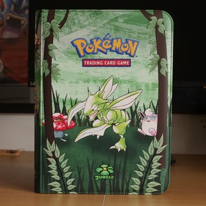 Op de afbeelding: Een rechthoekige Pokémon Trading Card Game map met een groene jungle-thema kaft. De voorkant toont het Pokémon-logo, het woord Jungle en illustraties van Scyther, Vileplume en Jigglypuff te midden van gestileerde bomen en bladeren.