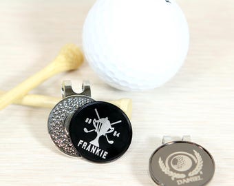 Puede incluir: Dos marcadores de bola de golf personalizados. Un marcador es negro con un diseño de trofeo plateado y palos de golf y el nombre Frankie. El otro marcador es plateado con un diseño de bola de golf y una corona de laurel y el nombre Daniel.
