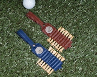 May include: Two leather golf tee holders, one brown and one blue, each holding wooden tees. A white golf ball rests on the green turf. The blue holder has the text "CHUCK 1971".