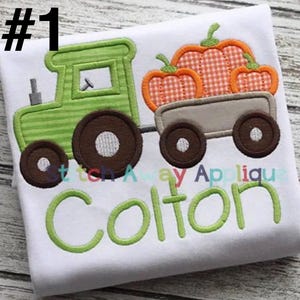May include: A white t-shirt featuring an embroidered applique of a green tractor pulling a gray wagon filled with three orange gingham pumpkins. The name Colton is embroidered in large green letters below the tractor. The number 1 is in the corner.