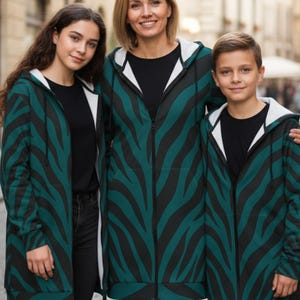 Peut inclure: Trois personnes portant des sweat-shirts &agrave; capuche zipp&eacute;s assortis, &agrave; rayures z&eacute;br&eacute;es teal et noires, avec des capuches doubl&eacute;es de blanc. Ils se tiennent &agrave; l'ext&eacute;rieur devant un arri&egrave;re-plan flou d'une rue de la ville. Les sweat-shirts sont longs, atteignant le milieu des cuisses.