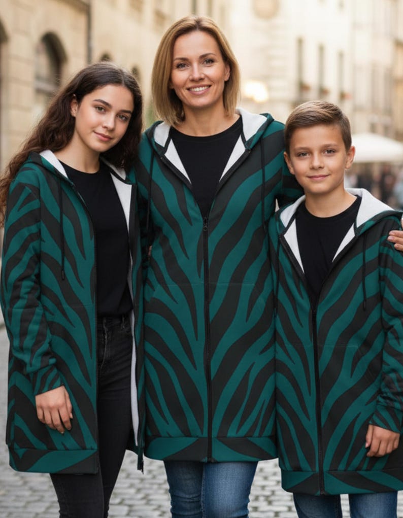 Peut inclure: Trois personnes portant des sweat-shirts &agrave; capuche zipp&eacute;s assortis, &agrave; rayures z&eacute;br&eacute;es teal et noires, avec des capuches doubl&eacute;es de blanc. Ils se tiennent &agrave; l'ext&eacute;rieur devant un arri&egrave;re-plan flou d'une rue de la ville. Les sweat-shirts sont longs, atteignant le milieu des cuisses.