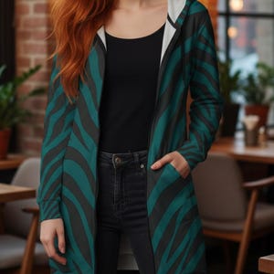 Peut inclure: Longue veste &agrave; capuche avec un motif z&eacute;br&eacute; teal et noir. La veste a une doublure int&eacute;rieure blanche et une fermeture &eacute;clair sur toute la longueur. La personne porte un haut noir et un jean noir. La veste a des poches.