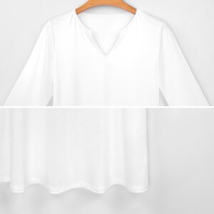 May include: A white long-sleeved shirt with a V-neck collar. The shirt is made of a soft, flowing fabric and hangs loosely. The hemline has a subtle wave pattern. The shirt is hanging on a wooden hanger.