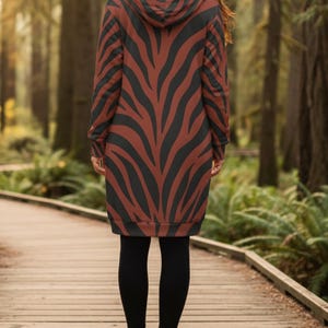 Peut inclure: Une longue robe &agrave; capuche avec un motif z&eacute;br&eacute; marron et noir. La robe est port&eacute;e avec des leggings noirs et des bottines noires. La personne se tient sur une passerelle en bois dans une for&ecirc;t.