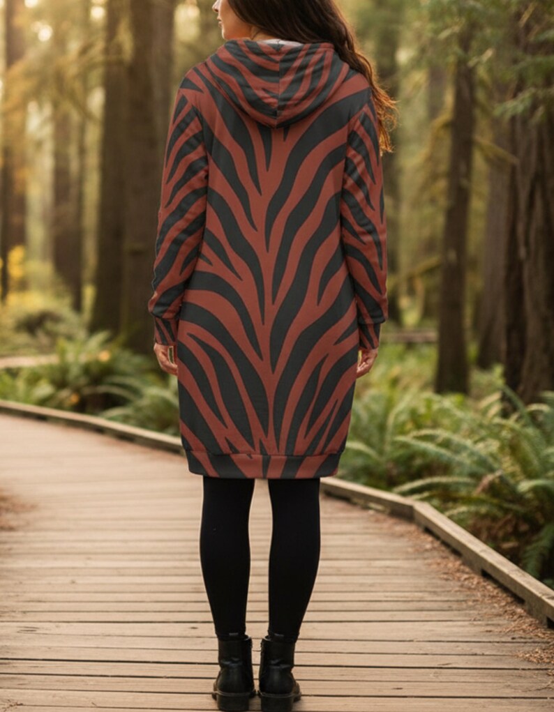 Peut inclure: Une longue robe &agrave; capuche avec un motif z&eacute;br&eacute; marron et noir. La robe est port&eacute;e avec des leggings noirs et des bottines noires. La personne se tient sur une passerelle en bois dans une for&ecirc;t.