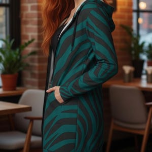 Peut inclure: Une longue veste &agrave; capuche avec un motif z&eacute;br&eacute; teal et noir. La veste a une fermeture &eacute;clair et des poches. L'int&eacute;rieur de la capuche est blanc. La personne porte un haut noir en dessous.